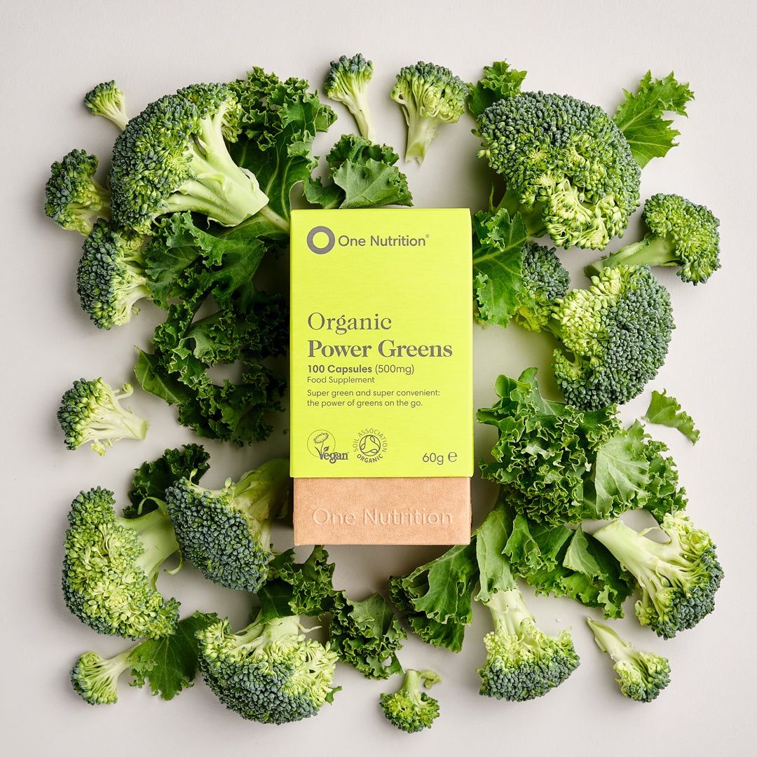 One Nutrition Organic Power Greens supplement package surrounded by fresh green vegetables on a light background