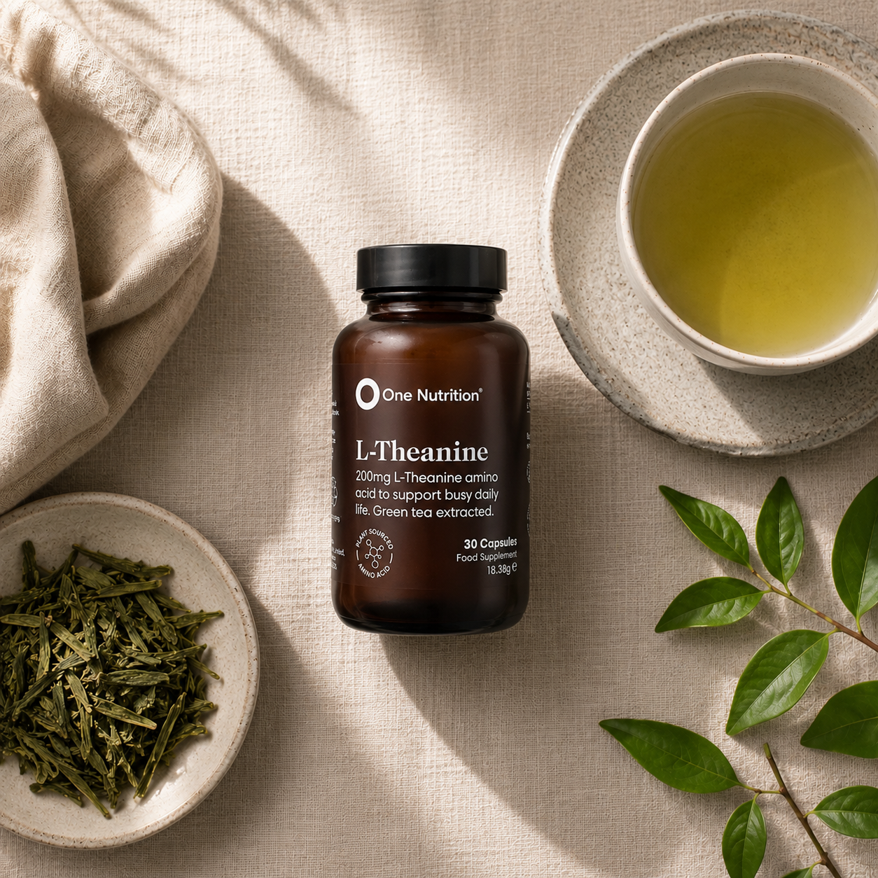 A bottle of One Nutrition L-Theanine 200mg dietary supplements, styled in a flat lay with a cup of green tea, loose green tea leaves, and green leaves on a beige linen background.