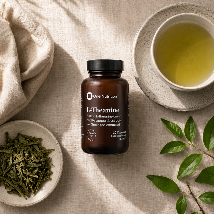 A bottle of One Nutrition L-Theanine 200mg dietary supplements, styled in a flat lay with a cup of green tea, loose green tea leaves, and green leaves on a beige linen background.